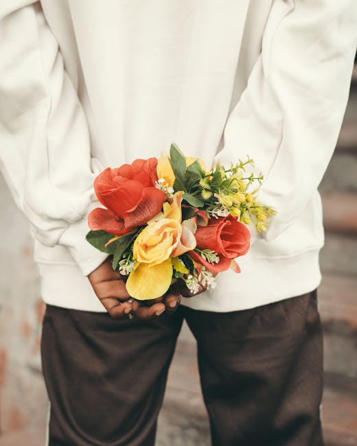 A person wearing a white jacket and dark trousers is holding a vibrant bouquet of fresh flowers behind their back. The bouquet features large coral and yellow roses, orange tulips, and smaller green a