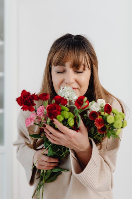 A woman with light brown hair and bangs, dressed in a beige sweater, gently holds a vibrant bouquet of fresh flowers close to her face with her eyes closed and a contented expression. The bouquet incl