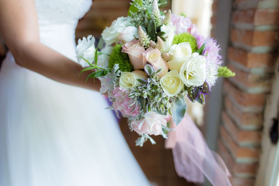 A bride holding a bouquet arrangement featuring cream and pale pink roses, white lisianthus, and light purple hydrangeas, complemented by green foliage and dusty miller. The bouquet is wrapped in soft