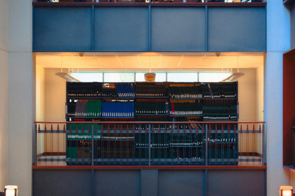 Photograph of a modern indoor space with a large bookshelf filled with black, blue, green, yellow, and red binders, positioned on a mezzanine level with a wooden railing. The bookshelf is centered in 