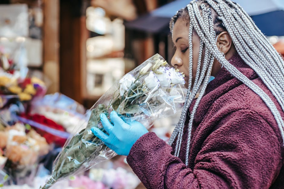 A woman with braided gray hair, wearing a maroon fleece jacket and blue gloves, is holding a large bouquet of white flowers wrapped in clear plastic at an outdoor flower market. The bouquet features f