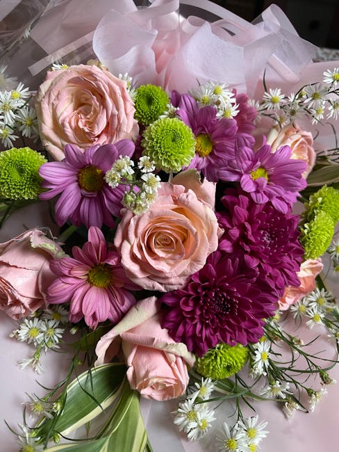 A vibrant floral bouquet featuring light peach roses, purple daisies, and bright green button chrysanthemums, accented with small white daisies and green foliage. The flowers are arranged in a balance