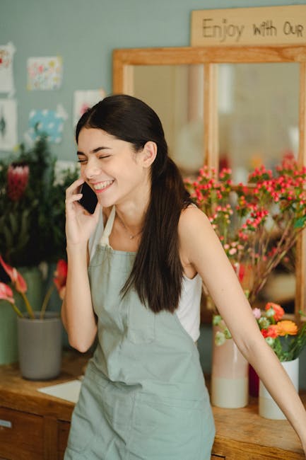 A young woman with long dark hair, smiling happily while talking on her mobile phone inside a floral shop. She is wearing a light grey apron over a white sleeveless top and is standing behind a wooden