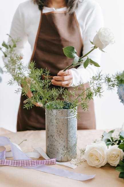 A floral arrangement process showing a person in a white shirt and brown apron carefully placing a single white rose with green stems and leaves into a silver, textured cylindrical vase. The designer'