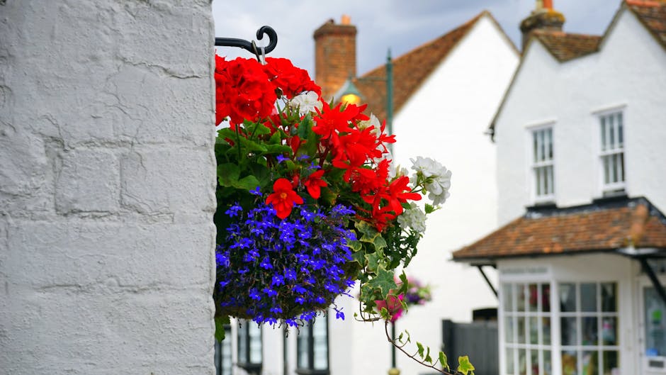 A vibrant hanging flower basket mounted on a black bracket attached to a white textured brick wall. The basket contains bright red geraniums, white petunias, and a profusion of deep blue lobelias, all