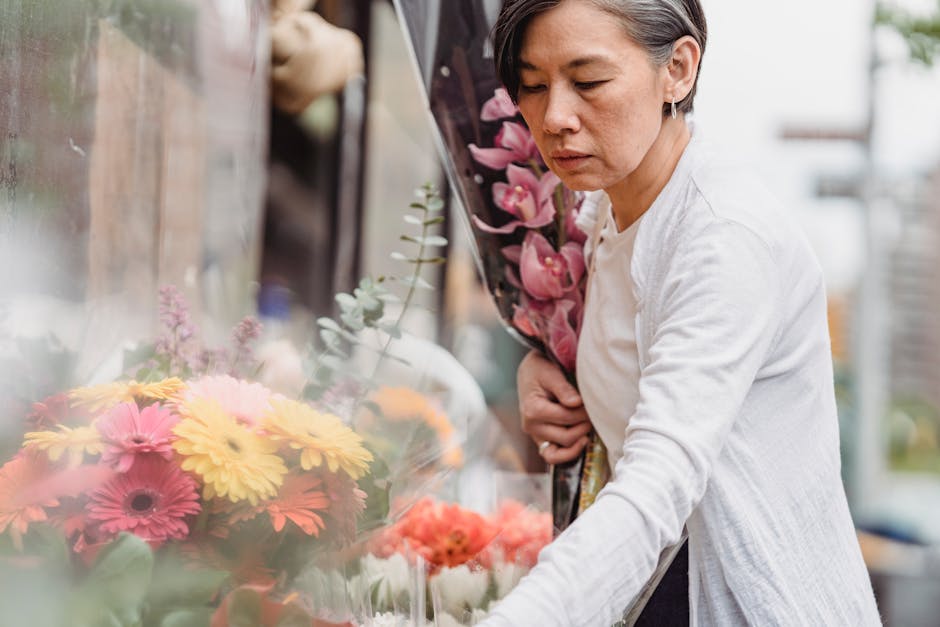 A mature woman with short dark hair, wearing a white cardigan, is selecting flowers at an outdoor flower shop. She holds a bouquet of pink and purple orchids against her chest while leaning slightly f