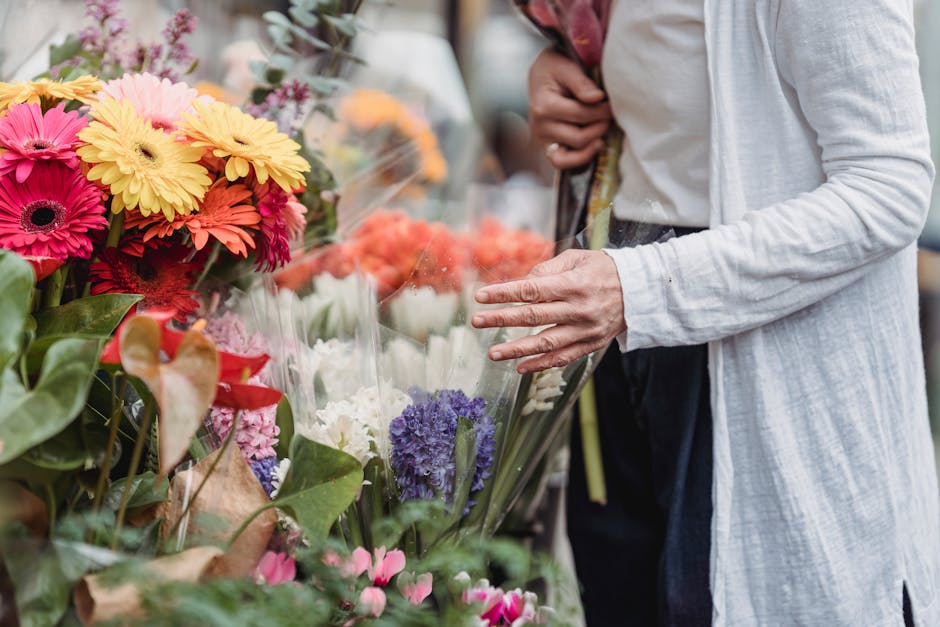 A person in a white long-sleeve shirt and dark trousers is selecting a bouquet of fresh flowers at a floral stall. The arrangement includes vibrant pink, yellow, and orange gerbera daisies, white carn