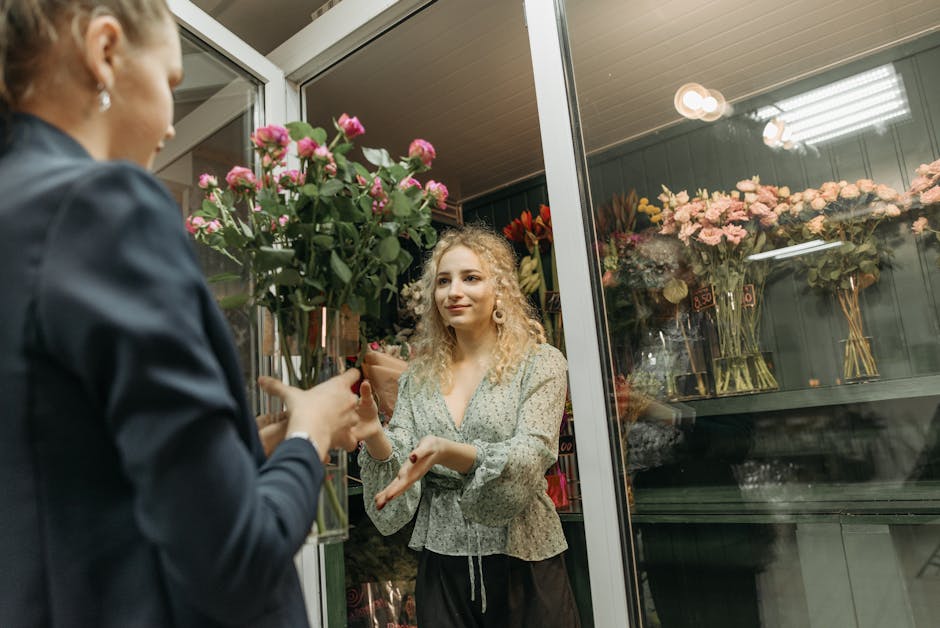 A woman with curly blonde hair, wearing a light-colored patterned blouse, is inside a floral shop, receiving a bouquet of pink roses wrapped in brown paper from a staff member dressed in dark clothing