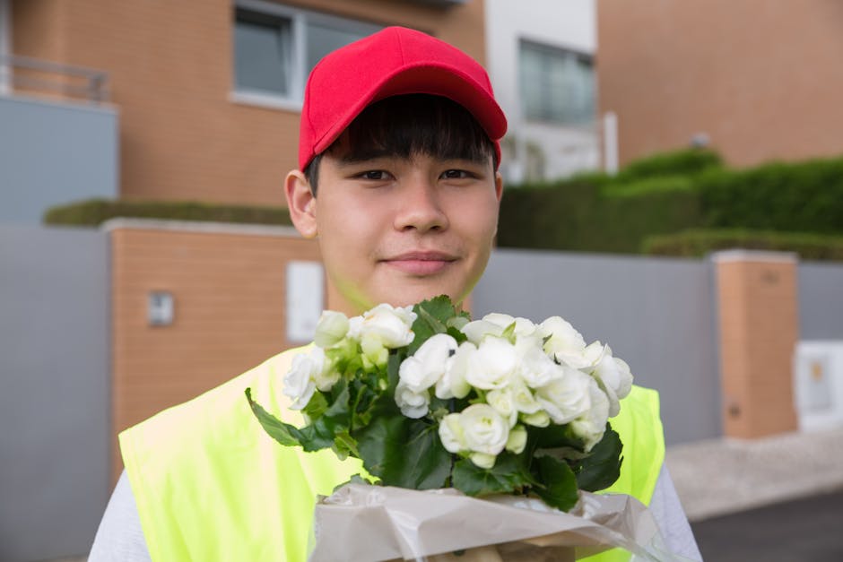 A young male flower delivery worker wearing a red cap and a bright yellow high-visibility vest is holding a bouquet of fresh white roses wrapped in white paper. He stands outdoors in a residential are